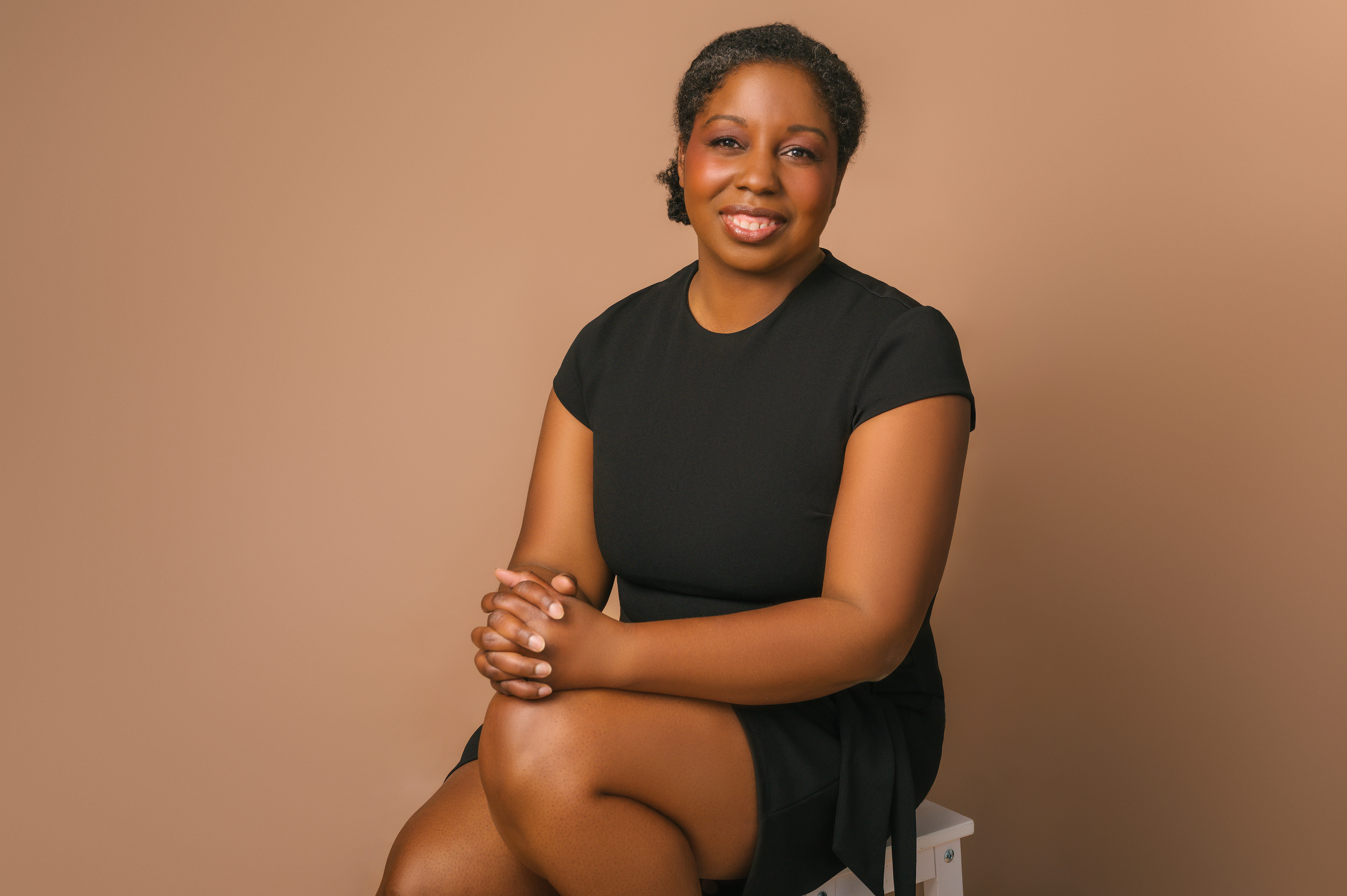 Tiffany faces the camera smiling. She is wearing a black dress and sitting on a stool.