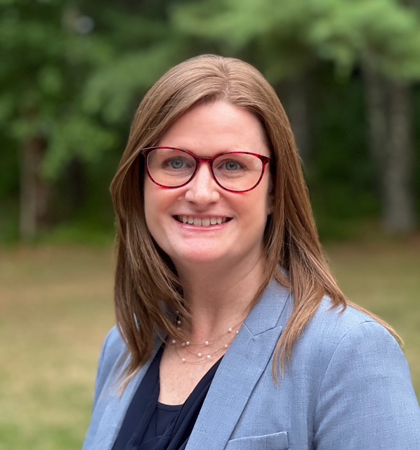 image of Kate with red rimmed glasses, blue suit coat in front of trees