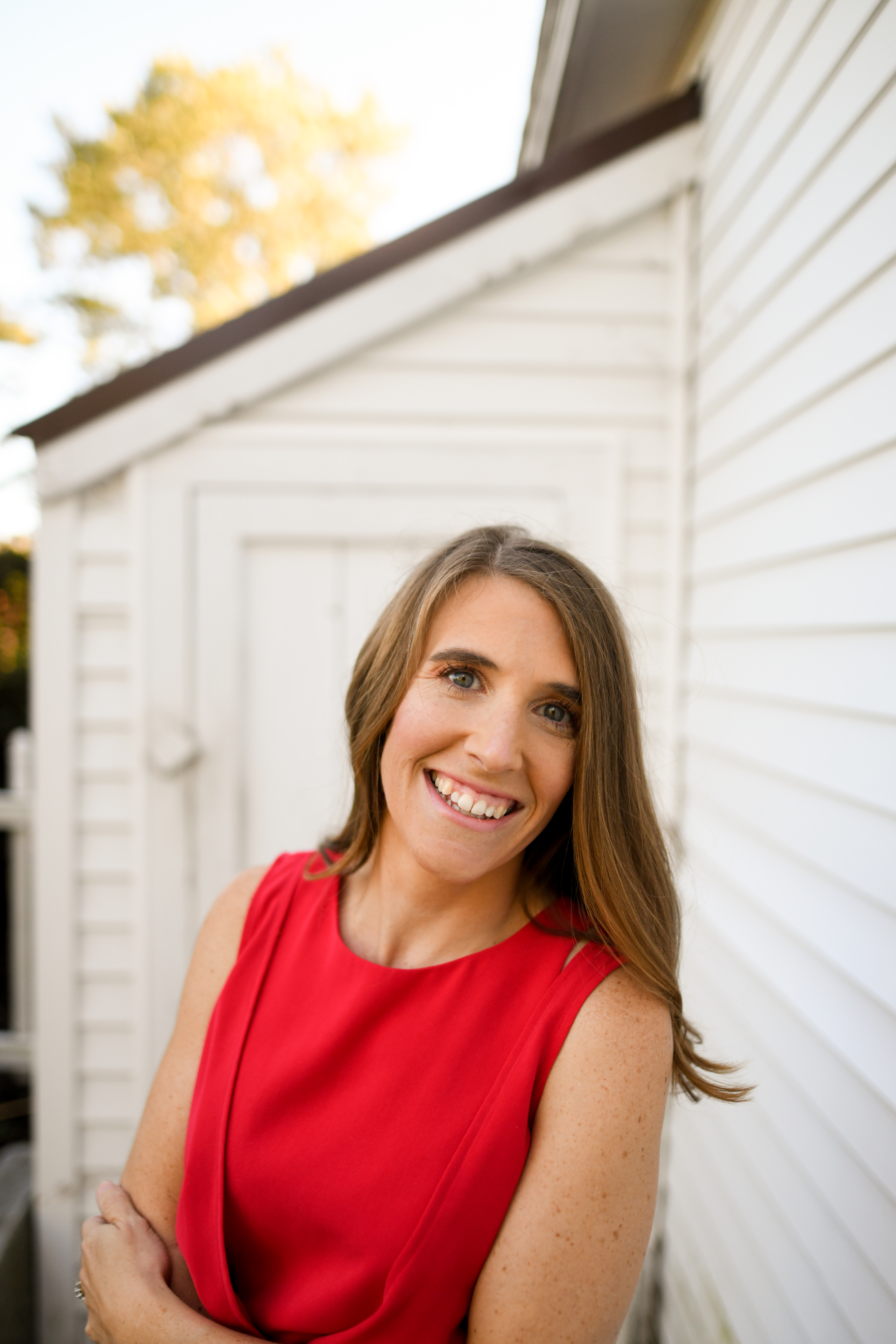 A portrait of a woman with shoulder-length brown hair, wearing a sleeveless red dress. She is smiling broadly at the camera. She is standing against the backdrop of a white building with a slightly visible rooftop. The background includes some greenery, possibly trees, with leaves that appear to be shifting color, indicating it may be autumn.