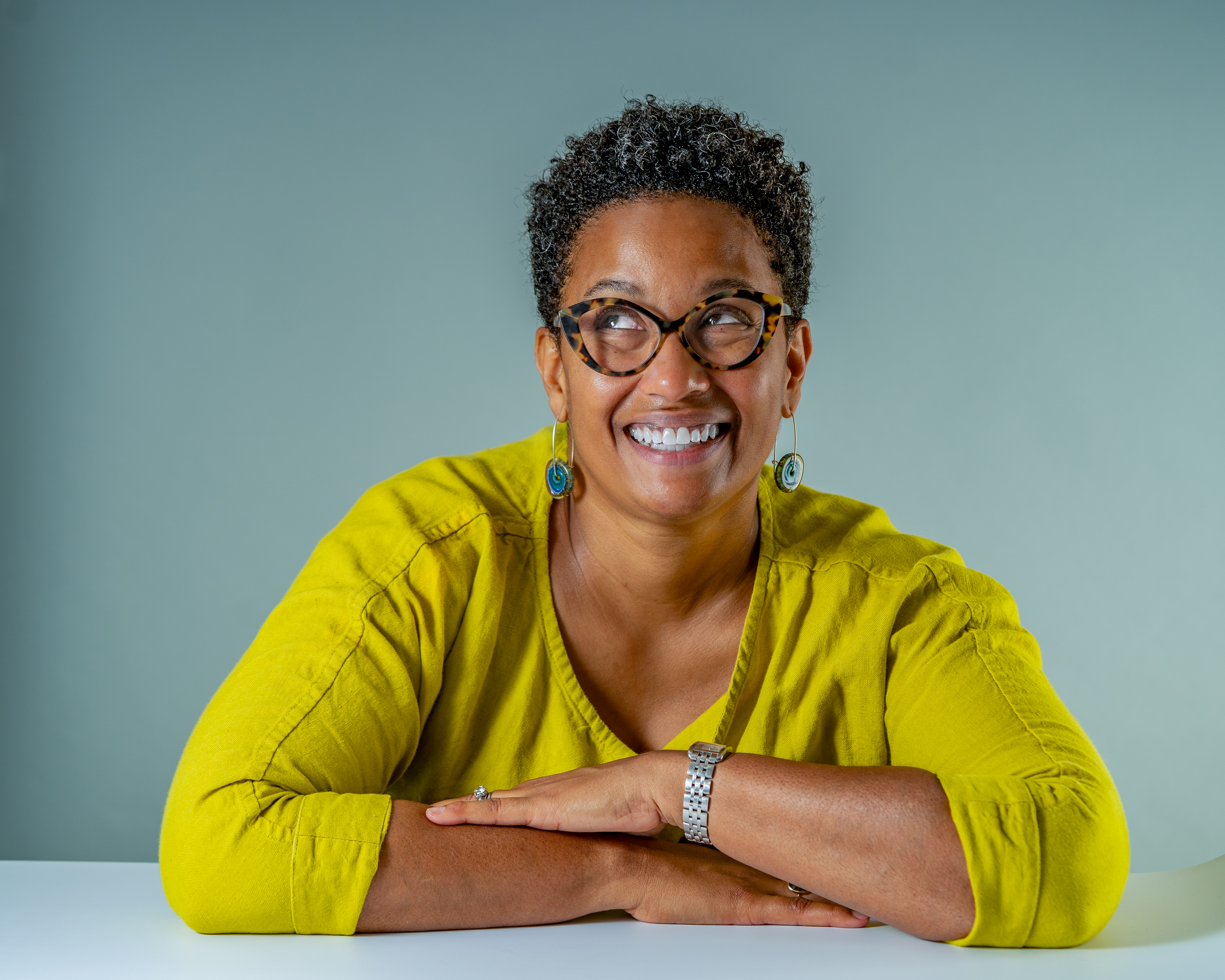 A light brown-skinned Black woman in a citron dress and large eyeglasses sits with her arms folded on a desk. She is smiling and looking upwards.