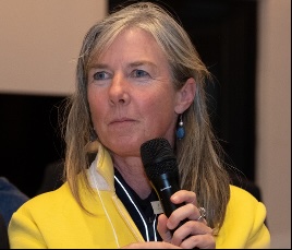 woman holding a mic  and wearing  a bright yellow jacket, listening to audience